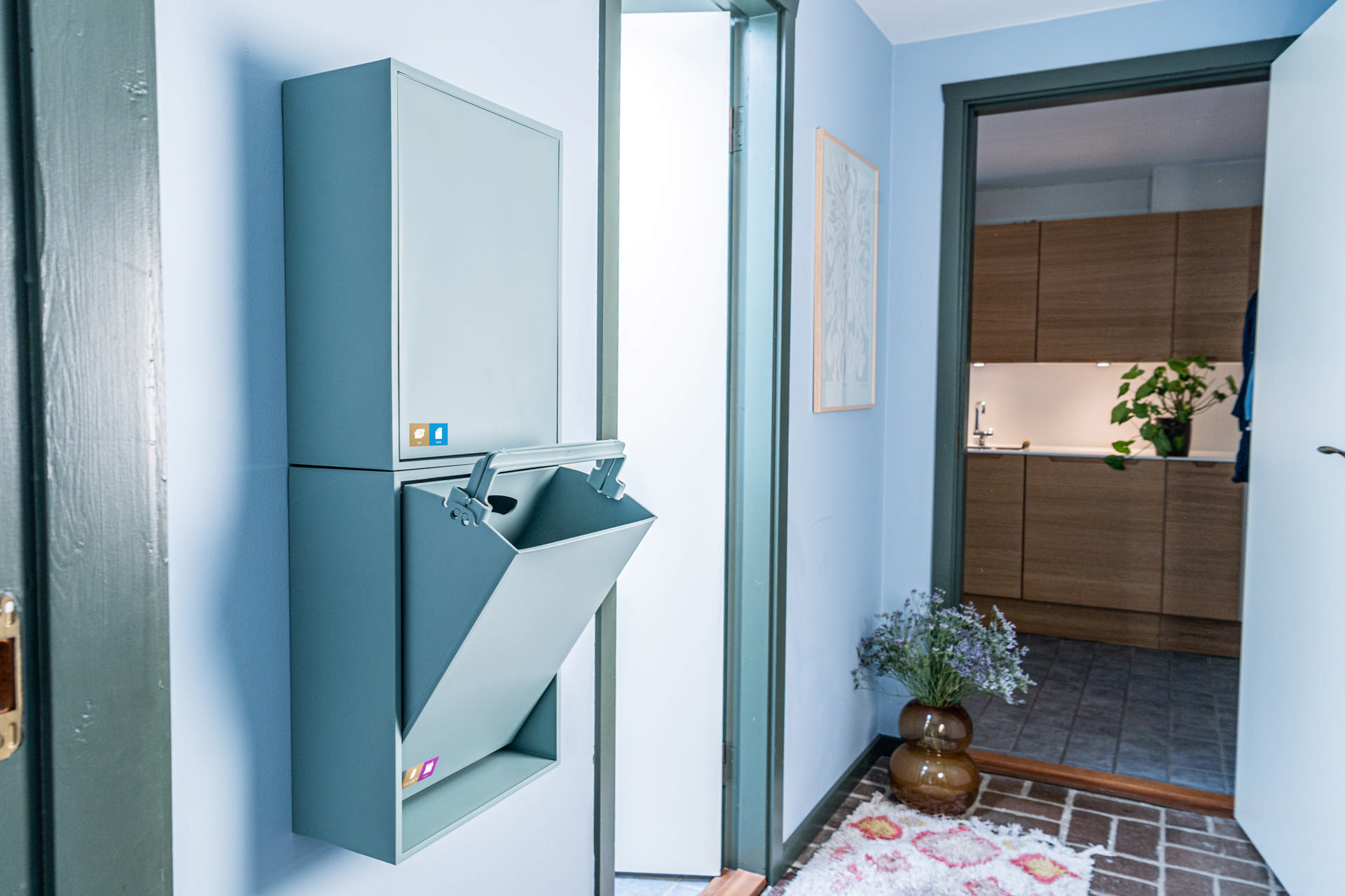 Smalle beholdere til papir/pap og metal/plast/mad- og drikkekartoner. Beholderne er opsat på en væg i et gangareal. Sorteringsbeholderne og farvetemaet i gangen er blåt. Klik på billedet for fuld størrelse.