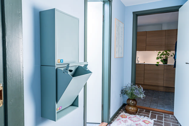 Smalle beholdere til papir/pap og metal/plast/mad- og drikkekartoner. Beholderne er opsat på en væg i et gangareal. Sorteringsbeholderne og farvetemaet i gangen er blåt. Klik på billedet for fuld størrelse.