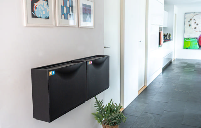 smalle beholdere til papir/pap og metal/plast/mad- og drikkekartoner. Beholderne er opsat på en væg i et gangareal. Beholderne er sorte, og væggen er hvid. Klik for at se i fuld størrelse.